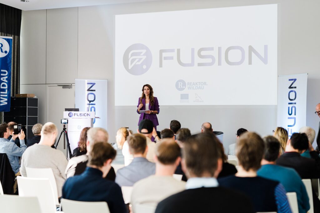 Moderatorin vor der Leinwand mit FUSION-Logo, Publikum von hinten, FUSION 2026 in Wildau