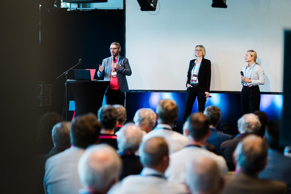 Drei Personen auf der Bühne beim Vortrag, Zuhörer von hinten, InnovationFestival Bielefeld 2026